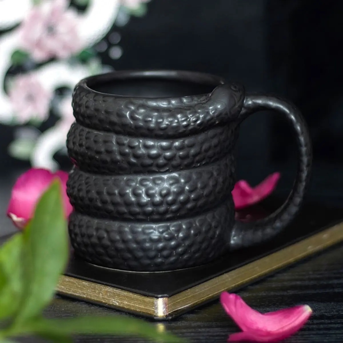 A black snake-shaped coffee mug with scale texture and rose petals on a golden-bordered surface.