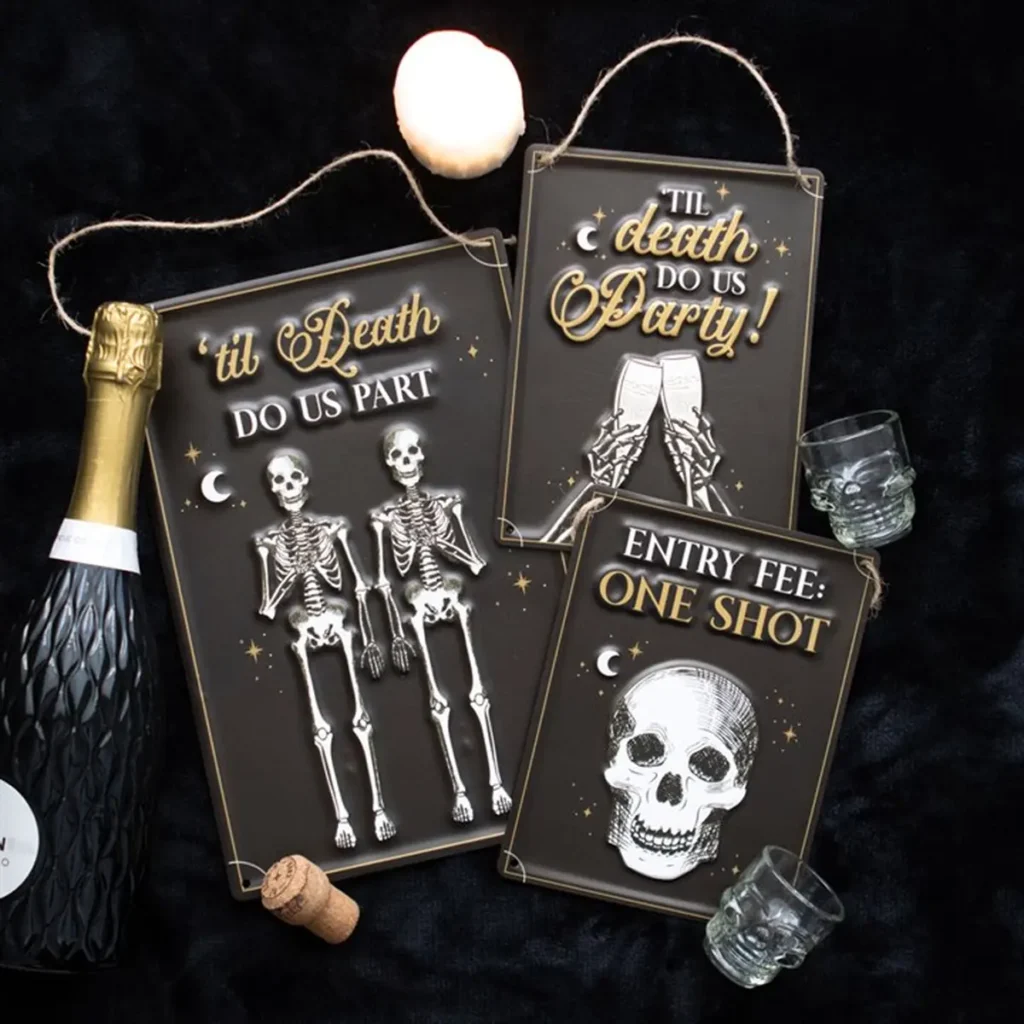 Halloween-themed skeleton party signs with gold lettering on a dark velvet surface, featuring champagne props.