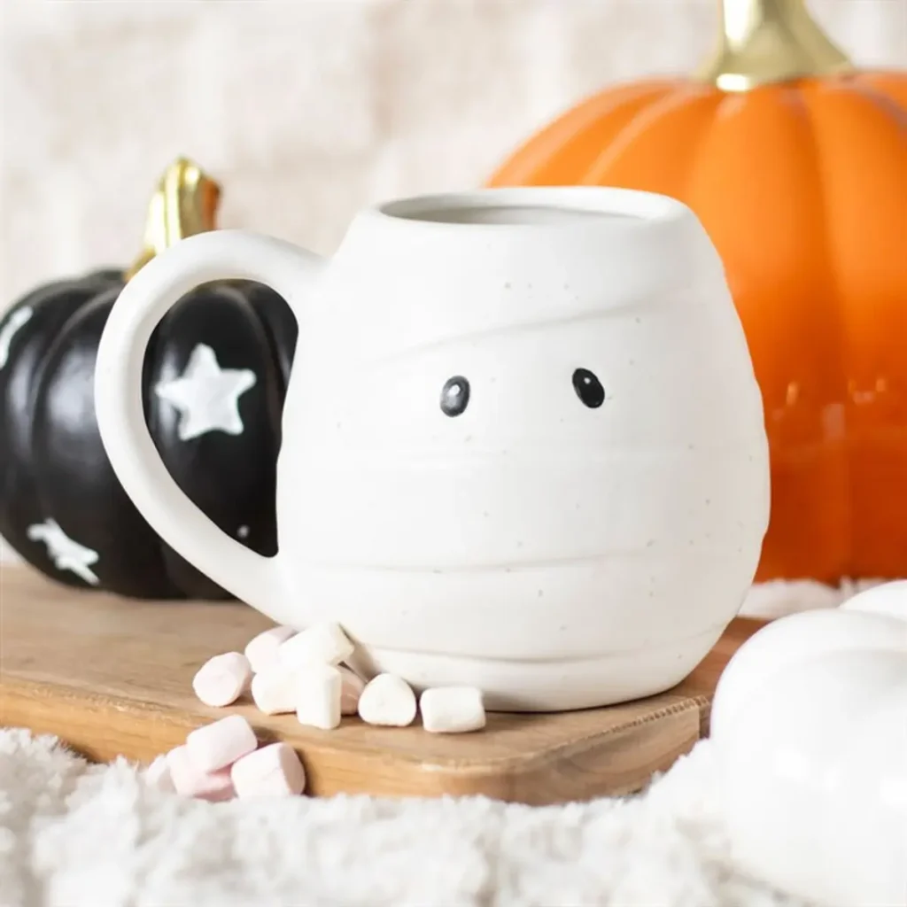 Halloween Decor Trends Worth Trying 9 Halloween Gift decor A white mummy mug with marshmallows and pumpkins on a cozy wooden surface.