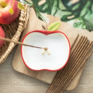 Apple Ceramic Incense Holder White Red - Natural Wooden Base A ceramic apple-shaped incense holder with incense sticks beside fresh red apples on a wooden surface.