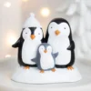 Penguin Family Figurine Resin Sculpture White Cozy Winter Theme A cute penguin family figurine on a snowy base with warm festive lights in the background.