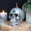 Silver Metallic Skull Sculpture With All-Seeing Eye Symbol A metallic skull with incense and all-seeing eye symbol on a wooden base beside a candle.
