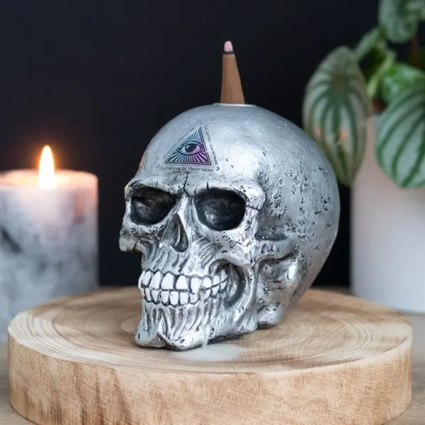 Silver Metallic Skull Sculpture With All-Seeing Eye Symbol A metallic skull with incense and all-seeing eye symbol on a wooden base beside a candle.