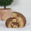 Hedgehog Family Sculpture Carved Wood Brown - Loving Embrace A wooden-style sculpture of three hedgehogs cuddling together beside a small potted green plant.