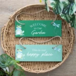 Two green wooden garden signs with frog and dragonfly designs inside a woven basket tray.