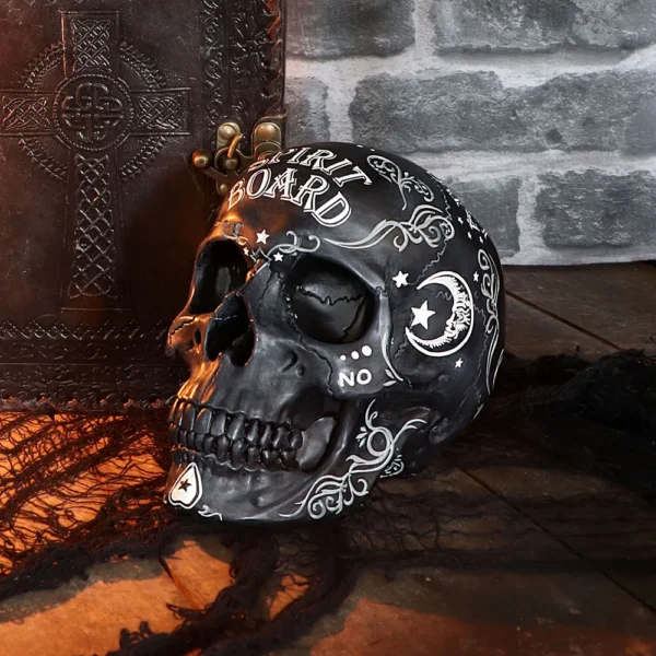 Black Ceramic Spirit Board Skull - Occult Design Decor A black decorative spirit board skull beside an embossed leather book on a wooden surface.