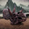 Triceratops Skull Fossil Sculpture - Reddish Brown Ancient Relic A weathered triceratops skull on reddish earth beneath misty mountains and an overcast sky.